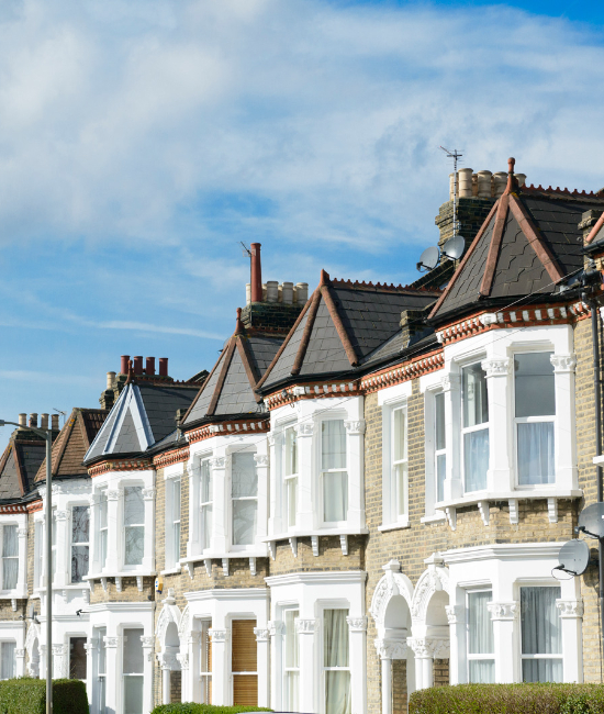 Row of traditional London terraced houses featuring yellow brick façades, white decorative window frames, ornate architectural details, and pitched roofs beneath a blue sky.